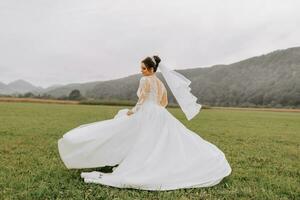 Bellissima, allegro sposa gioisce, balli, agitando sua vestito contro il sfondo di nuvole, alto nel il montagne nel un Aperto campo foto