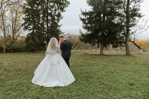 contento elegante coppia di Novelli sposi nel il verde foresta su un autunno giorno. il sposa nel un' classico lungo bianca vestito e il sposo nel un' blu completo da uomo abbraccio. nozze giorno. foto