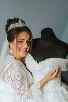 un' contento sposa è preparazione per sua lussuoso nozze a casa, Tenere sua bellissimo vestito nel sua mani. ritratto di un' donna con professionale capelli e trucco e un' Sorridi nel un' accappatoio foto