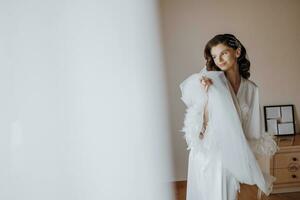 bellissimo giovane sposa con lungo Riccio capelli nel alla moda bianca accappatoio vicino finestra nel Hotel e Tenere velo nel mani. bellissimo giovane ragazza nel il mattina nel un' bianca vestire. gratuito spazio foto