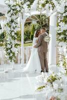 un elegante sposa e sposo posa insieme all'aperto su un' soleggiato nozze giorno contro un' sfondo di fiori nel un' bellissimo Posizione. il sposo delicatamente abbracci il sposa e baci suo. foto