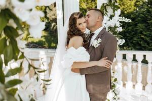 un elegante sposa e sposo posa insieme all'aperto su un' soleggiato nozze giorno contro un' sfondo di fiori nel un' bellissimo Posizione. il sposo delicatamente abbracci il sposa e baci suo. foto