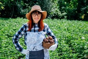 La contadina tiene una patata in un cesto di vimini che indossa un cappello di paglia e circondata da molte piante nel suo orto foto