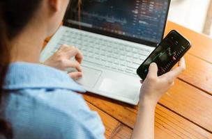 la mano della donna con il telefono e il computer portatile che controllano la borsa valori foto