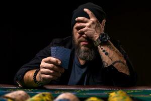 grave barbuto uomo seduta a poker tavolo e Tenere carte isolato su nero foto