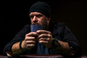 grave barbuto uomo seduta a poker tavolo e Tenere carte isolato su nero foto