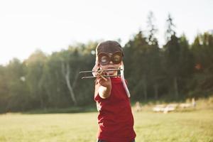 bambino felice nel casco pilota che gioca con un aeroplano giocattolo di legno e sogna di diventare volante foto