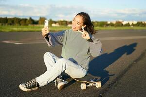 bellissimo coreano ragazza prende autoscatto su smartphone, prende foto con sua skateboard, enoys soleggiato giorno all'aperto