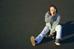 contento bellissimo coreano adolescente ragazza si siede su sua skateboard, crociera su longboard, indossare casuale Abiti foto