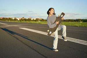 bellissimo asiatico adolescente ragazza giocando con sua longboard, Tenere skateboard come Se giocando chitarra, in piedi su strada su soleggiato giorno foto