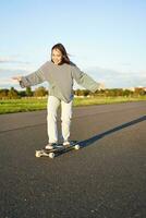 carino asiatico ragazza equitazione skateboard, pattinando su strada e sorridente. pattinatore su incrociatore longboard godendo all'aperto su soleggiato giorno foto