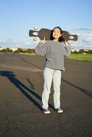 stile di vita e le persone. giovane asiatico ragazza in posa con longboard, pattinando su sua incrociatore. sorridente donna Tenere skateboard su le spalle foto