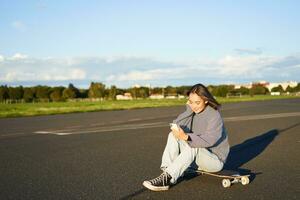 ritratto di giovane coreano ragazza seduta su sua skateboard su strada, guardare a smartphone, chat su mobile App foto