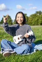 contento asiatico ragazza giocando ukulele nel parco, mostrando roccia n rotolo, pesante metallo corna cartello e sorridente, avendo divertimento all'aperto foto
