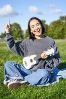contento asiatico ragazza giocando ukulele nel parco, mostrando roccia n rotolo, pesante metallo corna cartello e sorridente, avendo divertimento all'aperto foto