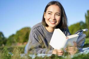 ritratto di carino coreano ragazza, lettura nel parco mentre dire bugie su erba, rilassante con preferito libro nel mani, sorridente felicemente foto