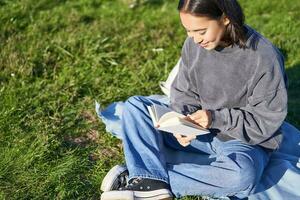 ritratto di asiatico ragazza lettura prenotare, seduta su sua coperta nel parco, con verde erba, sorridente felicemente foto