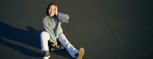 bellissimo coreano adolescente ragazza si siede su skateboard e gode soleggiato giorno. sorridente giovane asiatico donna pattinando su longboard foto