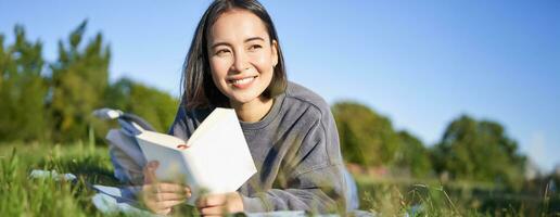 ritratto di carino coreano ragazza, lettura nel parco mentre dire bugie su erba, rilassante con preferito libro nel mani, sorridente felicemente foto