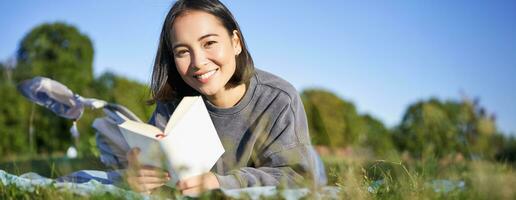 ritratto di carino coreano ragazza, lettura nel parco mentre dire bugie su erba, rilassante con preferito libro nel mani, sorridente felicemente foto