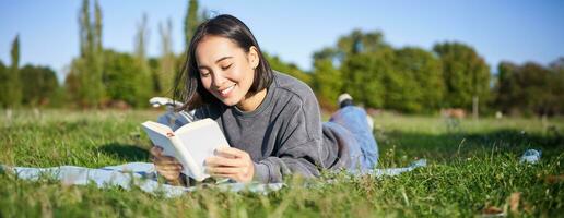 ritratto di bellissimo sorridente asiatico ragazza, lettura nel parco, dire bugie su erba con preferito libro. tempo libero e persone concetto foto