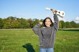 ottimista giovane donna danza con sua musicale strumento. ragazza solleva sua ukulele su e posa nel parco su verde campo foto