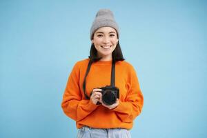 elegante asiatico ragazza con digitale telecamera, assunzione immagini. donna fotografo sorridente, in piedi al di sopra di blu sfondo foto
