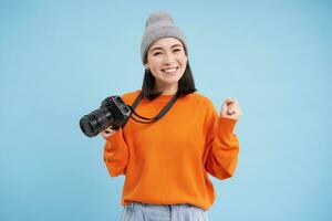 elegante asiatico ragazza con digitale telecamera, assunzione immagini. donna fotografo sorridente, in piedi al di sopra di blu sfondo foto