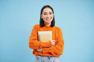 formazione scolastica e studenti. contento asiatico donna, Tenere i Quaderni e ridendo, sorridente a telecamera, gode andando per Università o Università, blu sfondo foto