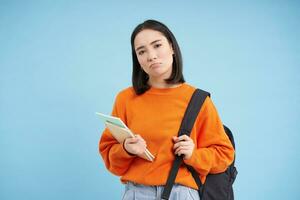 triste broncio asiatico alunno, ragazza con zaino e i Quaderni, guardare rovesciato, in piedi al di sopra di blu sfondo foto