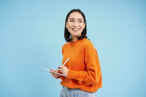 moderno bellissimo asiatico donna con digitale tavoletta e matita, assunzione Appunti, scrittura su sua gadget, fare compiti a casa, Lavorando, in piedi al di sopra di blu sfondo foto