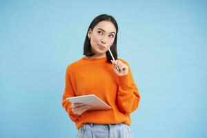 moderno bellissimo asiatico donna con digitale tavoletta e matita, assunzione Appunti, scrittura su sua gadget, fare compiti a casa, Lavorando, in piedi al di sopra di blu sfondo foto