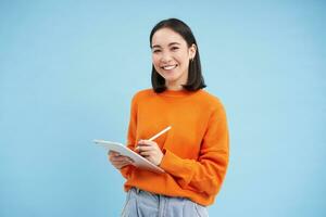 moderno bellissimo asiatico donna con digitale tavoletta e matita, assunzione Appunti, scrittura su sua gadget, fare compiti a casa, Lavorando, in piedi al di sopra di blu sfondo foto