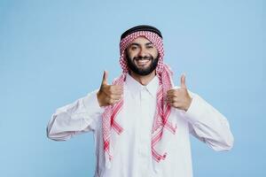 musulmano uomo indossare tradizionale arabo Abiti in piedi con pollici su gesto simboleggiante accordo e positività ritratto. allegro arabo vestito nel thobe e foulard mostrando approvare cartello foto