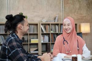 giovane bella donna medico sta esaminando la salute del paziente maschio in ufficio della clinica ospedaliera e consigliando con un sorriso sui farmaci. questo medico specialista asiatico è una persona islamica che indossa un hijab.