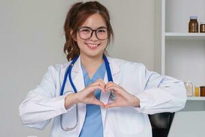 bellissimo asiatico donna indossare medico uniforme e stetoscopio sorridente nel amore fare cuore simbolo forma con mani. romantico concetto. foto