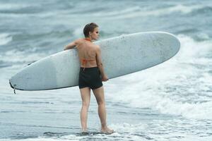 donna surfer a piedi nel caviglia in profondità acqua su spiaggia, trasporto tavola da surf e guardare a rottura onde foto