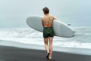 posteriore Visualizza di non identificato femmina surfer a piedi su nero sabbioso spiaggia mentre trasporto bianca tavola da surf foto