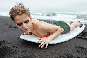 bionda surfer donna con luminosa trucco sembra sbalorditivo come lei bugie su tavola da surf su sabbioso spiaggia foto