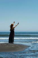donna nel lungo vestito in piedi su sabbioso spiaggia con sua braccia sollevato alto, raggiungendo su per cielo e mare foto