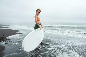 donna surfer a piedi nel caviglia in profondità acqua di mare onde su spiaggia, trasporto bianca tavola da surf foto