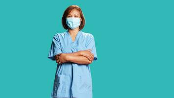 ottimista sorridente donna indossare protettivo clinico viso maschera per fermare batteri diffusione. ritratto di vivace infermiera nel professionale scrub con braccia piegato, isolato al di sopra di blu studio sfondo foto