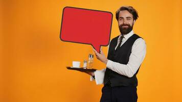 elegante Cameriere detiene discorso bolla icona su telecamera, Lavorando nel ristorante industria per creare marketing anno Domini con rosso cartone ritagliare. dipendente presentazione tabellone con vuoto isolato spazio. foto