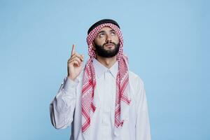 musulmano uomo vestito nel tradizionale Abiti puntamento verso l'alto con dito. giovane arabo indossare islamico thobe e scacchi copricapo mostrando e guardare su mentre in posa nel studio foto