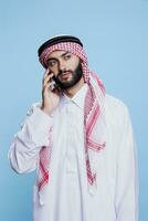 musulmano uomo indossare tradizionale thobe e foulard guardare lontano mentre A proposito di su smartphone. arabo parlando su mobile Telefono con neutro espressione mentre in piedi nel studio su blu sfondo foto