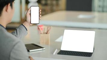 il giovane uomo d'affari tiene in mano uno smartphone con schermo vuoto e un laptop con schermo vuoto sulla scrivania.