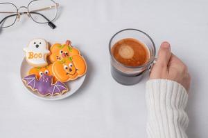 mano della donna che tiene la tazza di caffè durante il consumo di biscotti divertenti di halloween. buon giorno di halloween, trucco o minaccia, ciao ottobre, autunno autunno, tradizionale, festa e concetto di vacanza foto