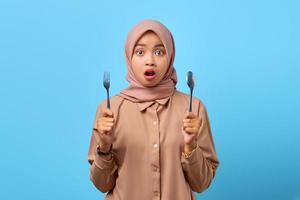 ritratto di giovane donna asiatica sorpresa tenere forchetta e cucchiaio con la bocca aperta su sfondo blu