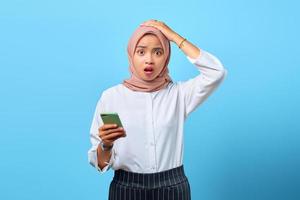 ritratto di giovane donna asiatica sorpresa che usa il telefono cellulare con la mano sulla testa su sfondo blu