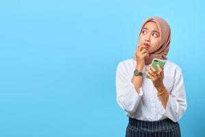 ritratto di giovane donna asiatica che pensa a qualcosa e tiene il telefono cellulare su sfondo blu foto
